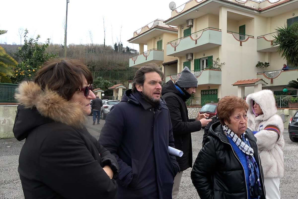 un gruppo di persone, in piedi nel mezzo di un piazzale, di fronte a delle abitazioni