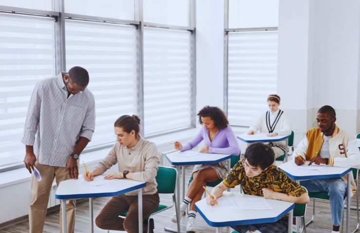 dei ragazzi seduti al bianco durante un compito e il maestro in piedi