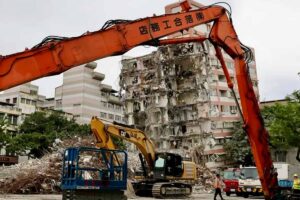 Il braccio di una ruspa e, sullo sfondo, un edificio danneggiato dal terremoto