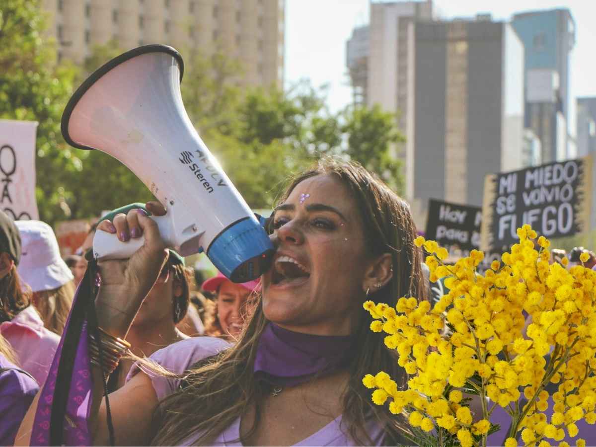 Una giovane donna con un megafono in mano urla i suoi diritti nel giorno dell'8 marzo. In primo piano, un mazzo di mimose, simbolo della Giornata della Donna