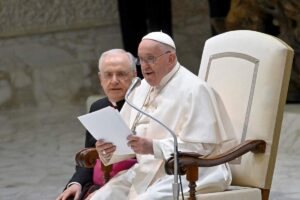 papa francesco seduto legge un foglio a un microfono e vicino a lui un vescovo