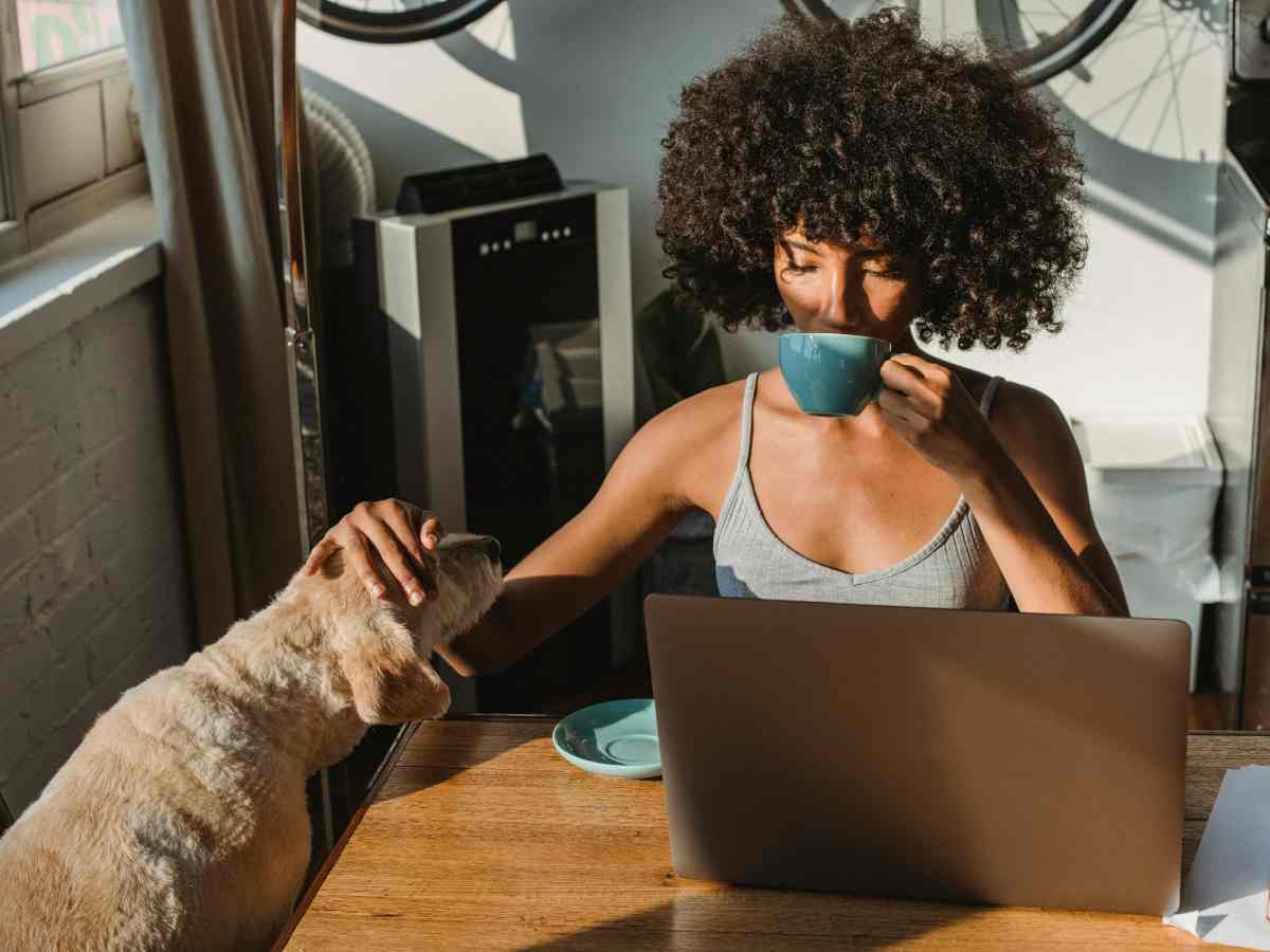 Ragazza che beve un caffè e accarezza un cane, seduta davanti al computer